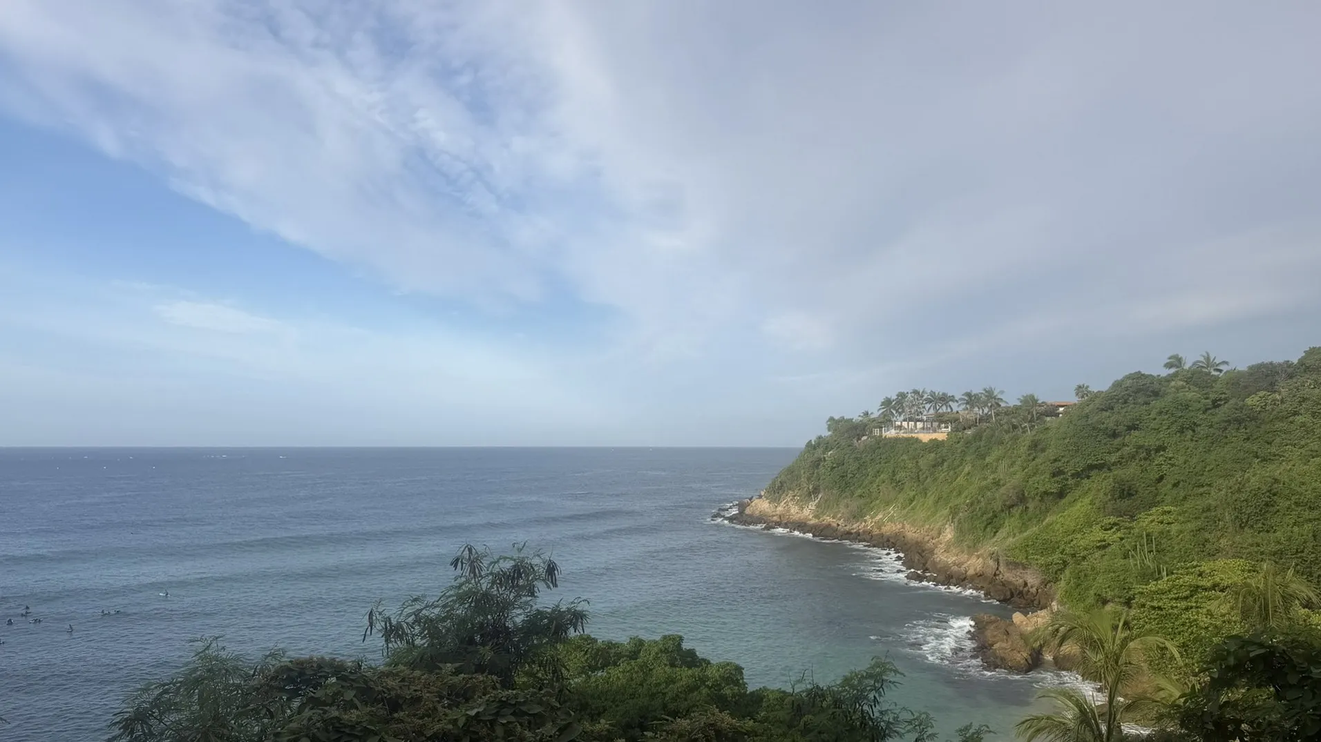 A bay on the coast of Puerto Escondido, with the Pacific, a rocky point and green hills under a wide sky